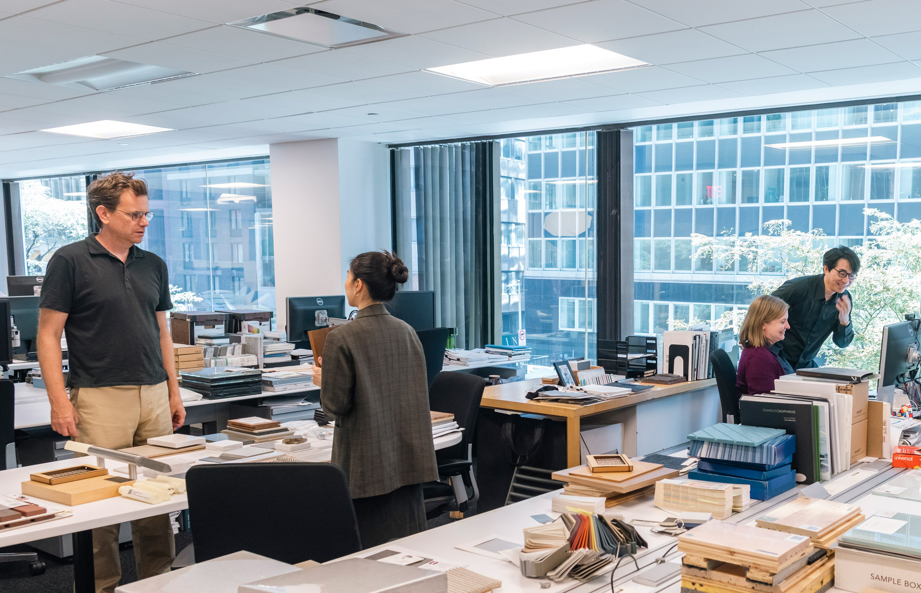 Open-plan architecture studio with staff conversing at workstations surrounded by material samples, sample boxes, and floor-to-ceiling city views.
